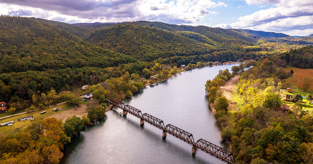 This is an image of a river with a trail bridge surrounded by mountains with fall foliage.