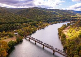 This is an image of a river with a trail bridge surrounded by mountains with fall foliage.