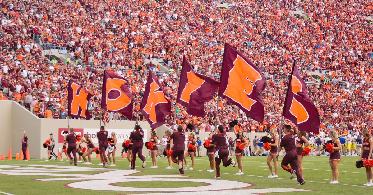 Hokie cheerleaders run onto the field with flags that spell ‘HOKIES.’