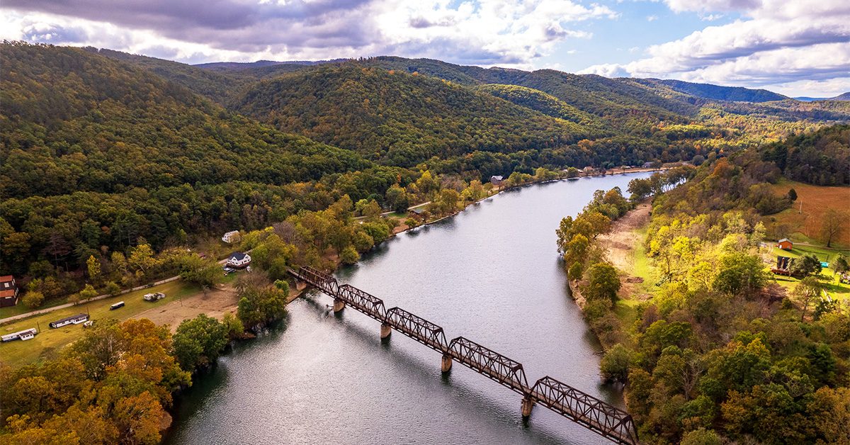 This is an image of a river with a trail bridge surrounded by mountains with fall foliage.